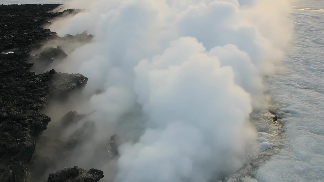 Pennacchio di vapore bianco che sale, costa vulcanica che emette vapore mentre il mare entra in contatto con la roccia calda, onde spumeggianti.