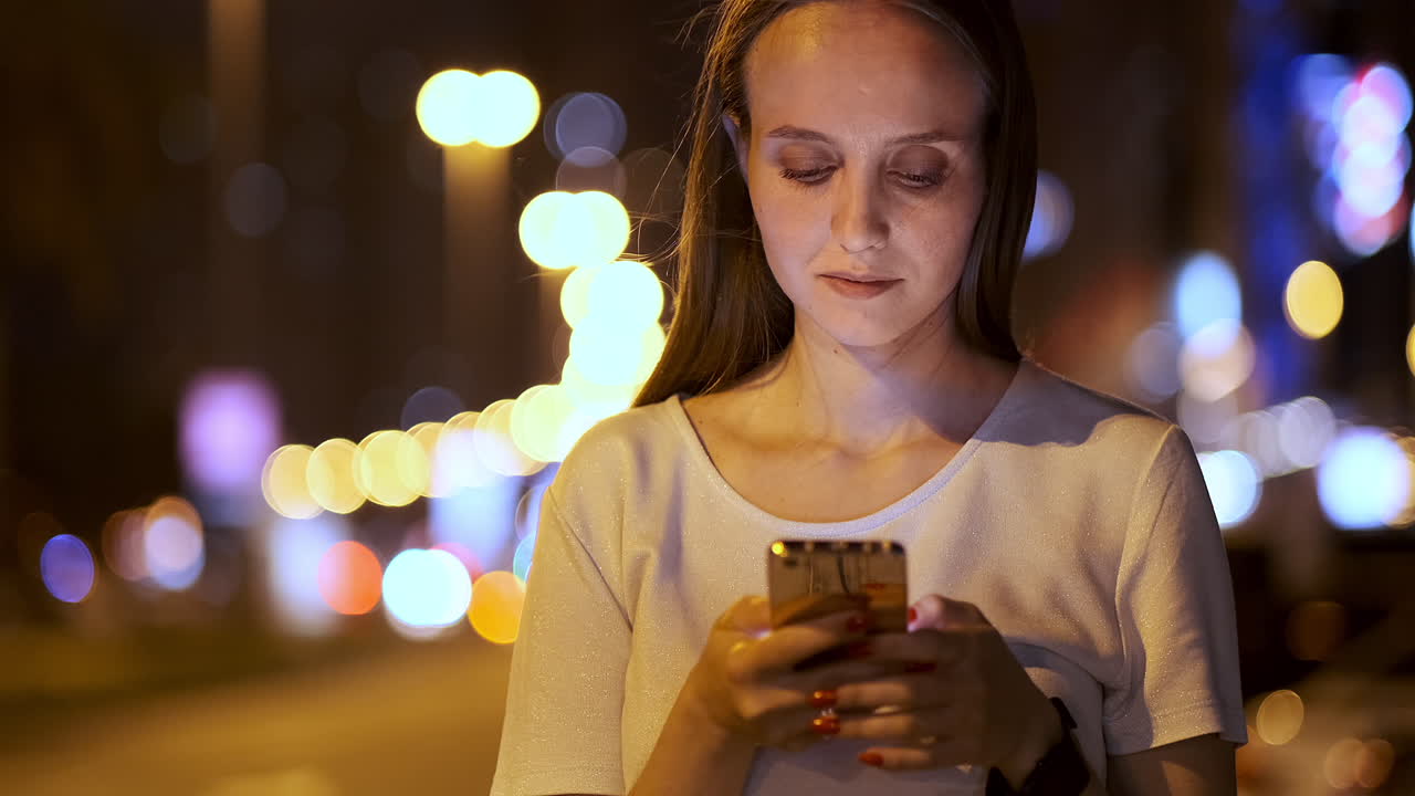 A beautiful young girl in the summer in a big night city keeps a smart phone in shell and writes a message reads e-mail and communicates in chat rooms and social networks keeps a blog looks at the phone screen