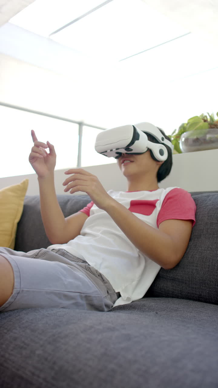 Vertical video: Using VR headset, Asian teenage boy sitting on couch, experiencing virtual reality a