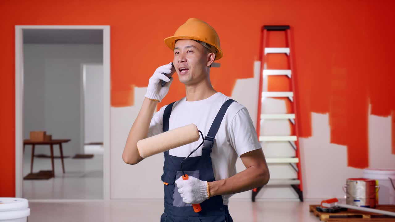 Side View Of Asian Man Painter Wearing Safety Helmet Talking On Smartphone And Looking Around While Standing With Painting Wall In Room