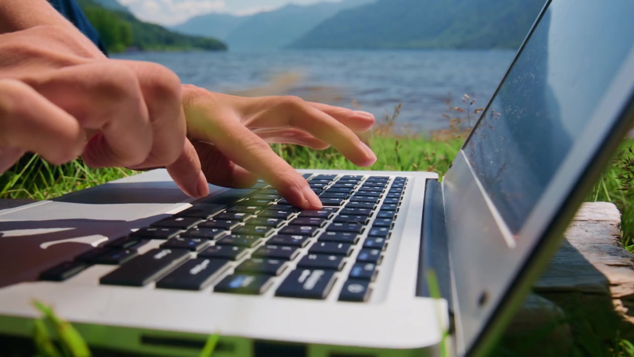 Hands type on laptop near lake. Green grass surrounds device. Mountains in background. Peaceful, focused outdoor work vibe. Ideal for remote work, digital nomad, travel, or nature-themed projects