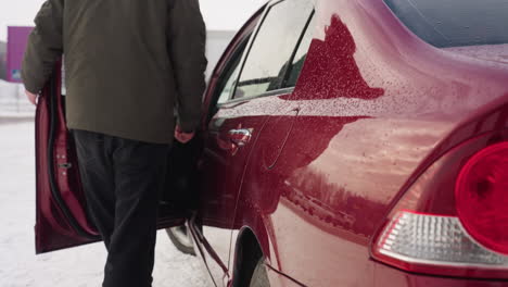 young man in winter jacket approaches parked red car on snowy ground with clear reflections of buildings, cars, and self on glossy surface, while snow lightly falls in urban industrial background