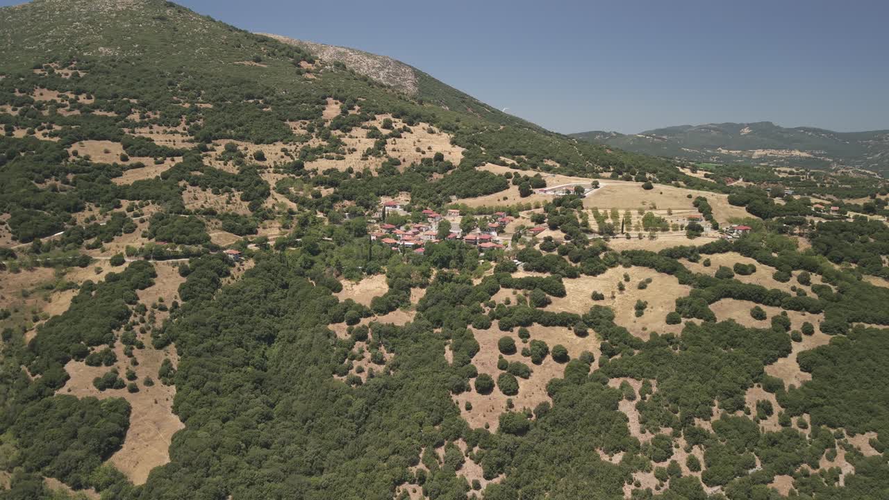 Ripresa aerea con drone in allontanamento che rivela un tradizionale villaggio greco a Mikros Pontias, circondato da un suggestivo paesaggio montano in una giornata di sole.