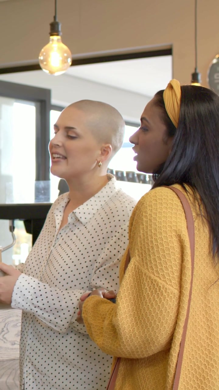 Vídeo vertical: Mulher careca de camisa de bolinhas se inclinando para fazer um pedido no café, enquanto um cardigã amarelo pega uma xícara.