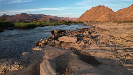 aerial drone flies past a man on a rocky riverbed during golden hour, shading his eyes as he gazes into the sunlit distance beneath warm, glowing skies