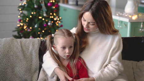 mother and daughter sitting on the sofa covered by a blanket while talking in living room with christmas decorations 1