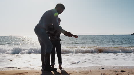 father teaching son to skim stones as they walk along beach by breaking waves together