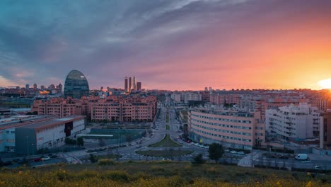 Timelapse-skyline-of-modern-european-city-Madrid-from-Las-Tablas-viewpoint-during-sunset-with-golden-light-and-red-clouds-during-blue-hour-zoom-in-day-to-night-time-lapse-yellow-flowers-foreground