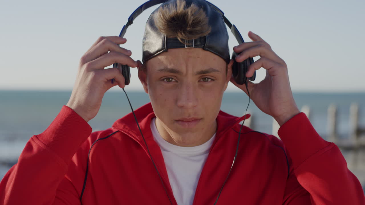 retrato de un adolescente confiado que se pone los auriculares escuchando música disfrutando de un relajado día soleado en la playa llevando la moda de moda en cámara lenta