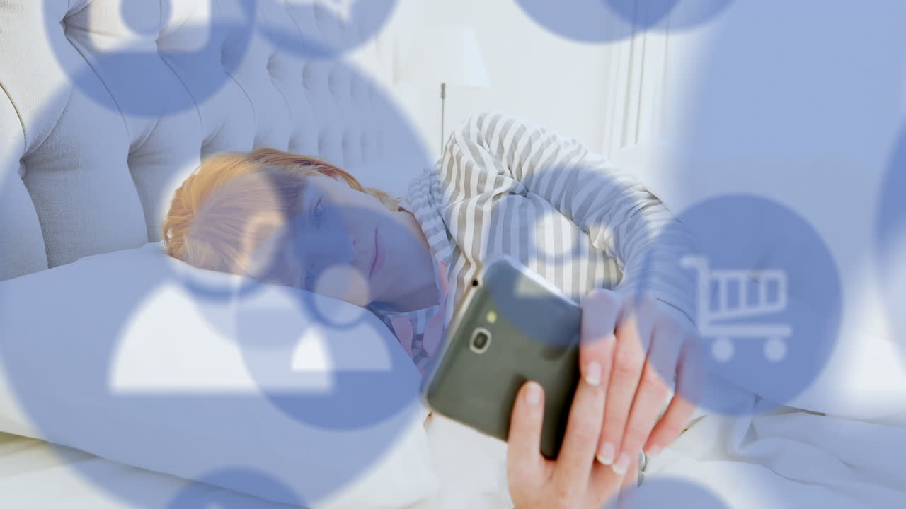Animation of multiple digital icons over caucasian woman using smartphone lying on the bed at home