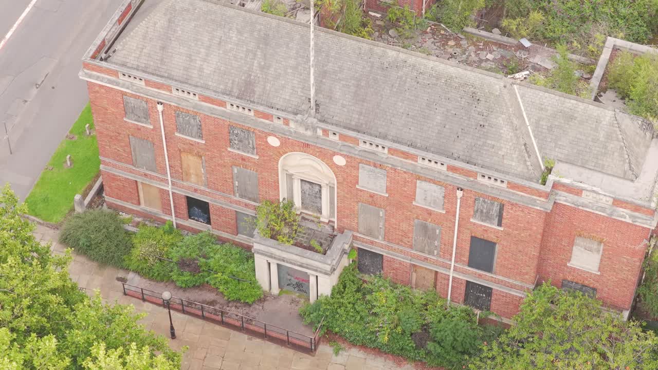 Drone ascending past the boarded-up façade of the abandoned Salford Crescent Police Station, highlighting its decayed brickwork, overgrown doorway, and silent reminder of lost civic use