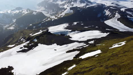 Aerial-shot-of-mountains-with-patches-of-snow-and-glacier-in-a-distance-partially-covered-by-white-clouds