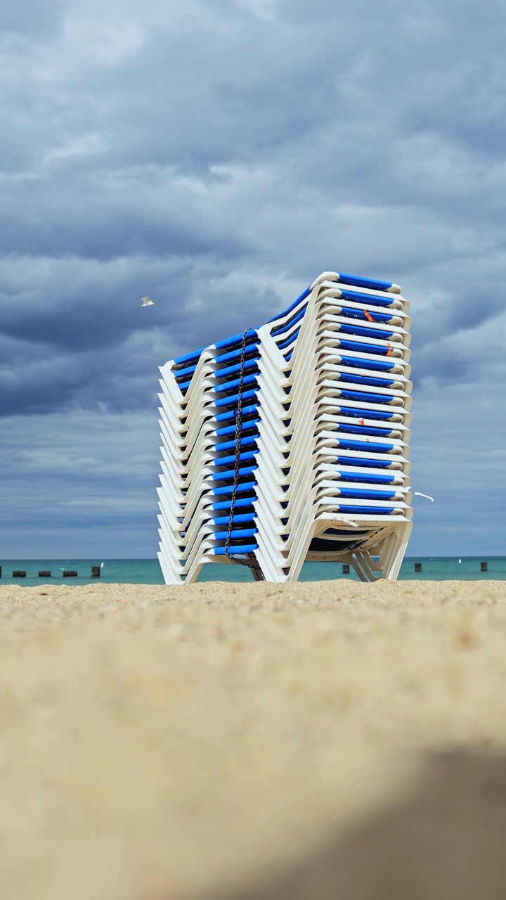Espreguiçadeiras empilhadas em uma praia de areia junto ao mar sob um céu nublado à tarde.