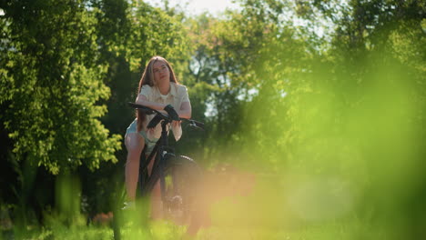 lower angle view of lady leaning on bike handlebar, gazing contemplatively with a subtle smile, sunlight gently reflects off her, background is filled with vibrant greenery