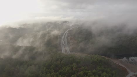 Drone-footage-of-highway,-and-housing-estate-in-Norway-with-some-fog-floating-in-the-air-and-sun-lighting-up-the-clouds