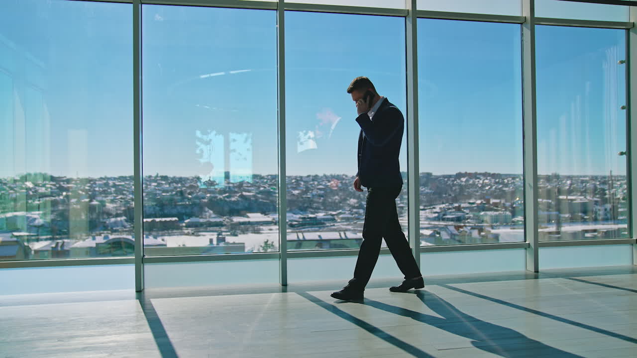 Full-length portrait of businessman. Male entrepreneur in elegant suits walking in modern office and talking about business at his smartphone. Panoramic windows view on city