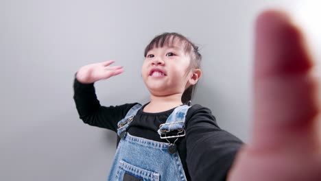 Happy-little-Asian-girl-using-a-smartphone-for-video-call,-waving-hand-and-speaking-and-talking-with-a-smile-greeting-teacher-and-classmates