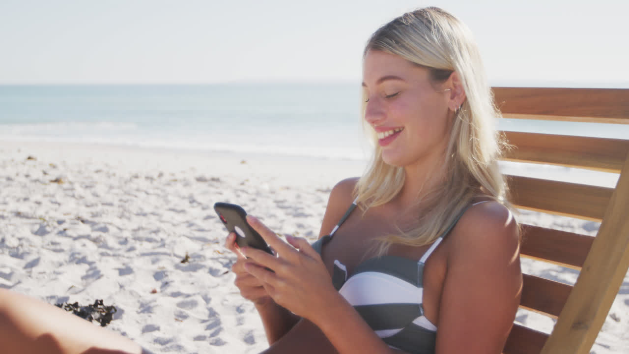 카우카스 여성이 해변에서 자리에 앉아 스마트폰을 사용하고 있습니다.