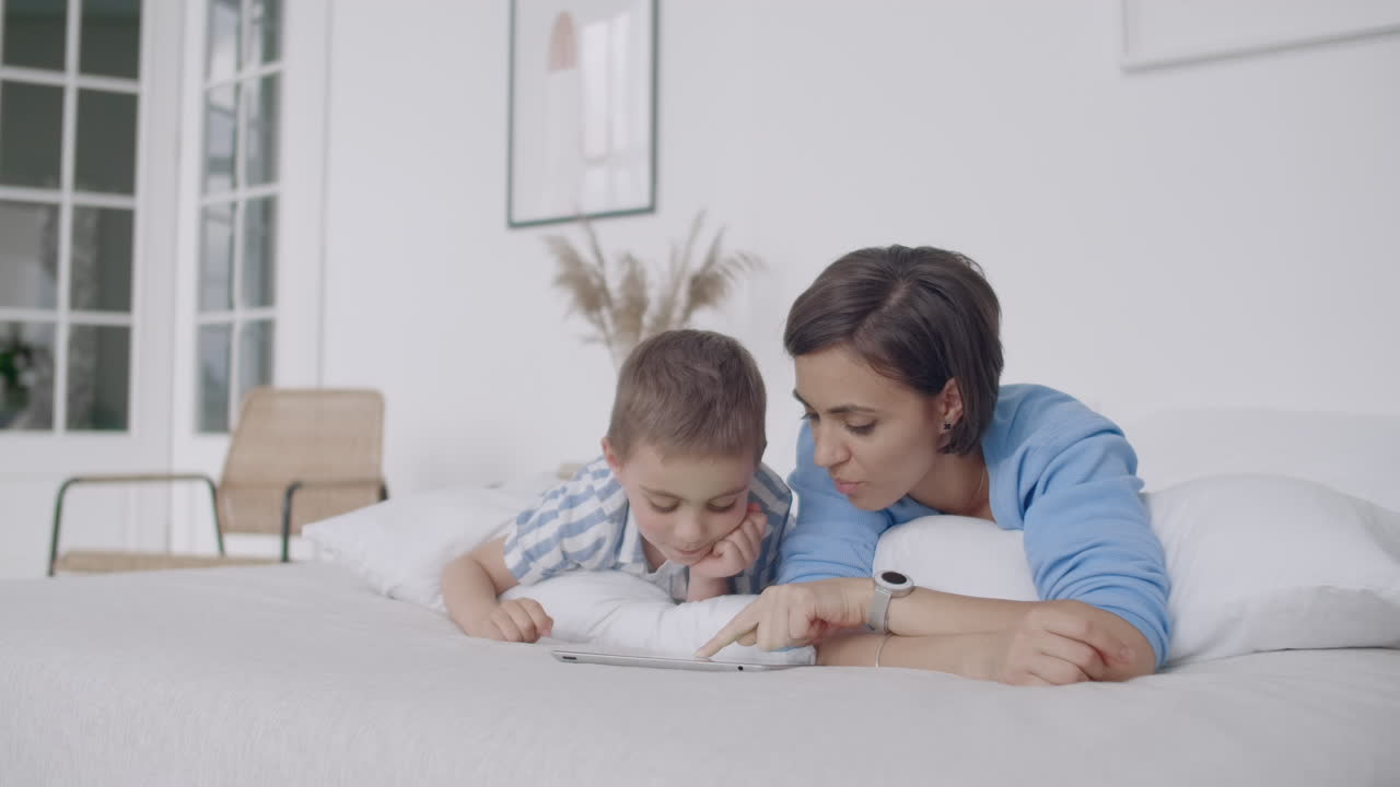 en el dormitorio blanco madre e hijo miran la pantalla de la tableta y se ríen. familia feliz en la cama por la mañana leyendo un libro y haciendo clic en el touchpad de la computadora.