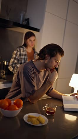 Vertical-video-scene-setting-of-a-brunette-man-in-a-gray-T-shirt-reading-a-book-after-dinner-while-his-brunette-girlfriend-in-a-checkered-shirt-plays-with-a-kitchen-knife-in-a-modern-apartment.-Scene-from-horror-films-a-brunette-girl-plays-with-a-knife-near-her-boyfriend