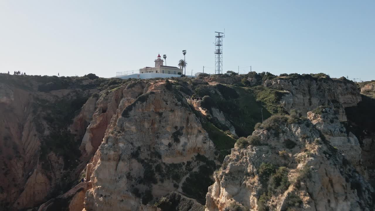 Aerial drone moves away from Ponta da Piedade, flying through dramatic limestone cliffs, rugged rock formations, and coastal greenery with the iconic lighthouse visible on the cliff top