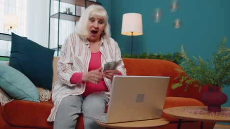 cheerful satisfied elderly grandmother freelancer woman pressing button on laptop and counting money