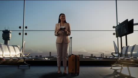 full body of asian businesswoman with rolling suitcase in boarding lounge at the airport, waiting for flight looks at passport then looking around, airplane takes off outside the window