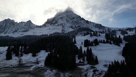 Dark-pine-trees-at-edge-of-dirt-at-base-of-backlit-snow-and-cloud-covered-mountain-peak