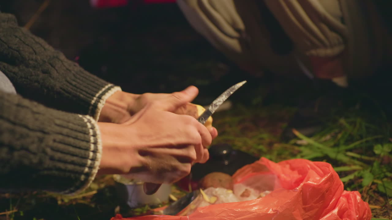 Close up of individual peeling potato with small knife during night camping on forest ground, hands focused on food preparation beside plastic bag and canned ingredients under warm light