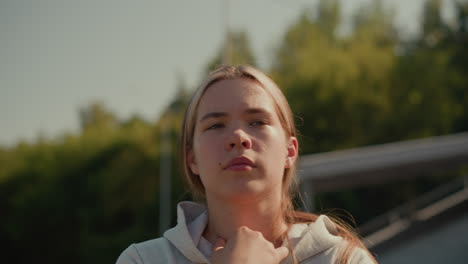 close-up of a thoughtful young woman walking in an outdoor stadium with her hand resting on her hoodie, looking pensive, the blurred background features green trees and electric poles