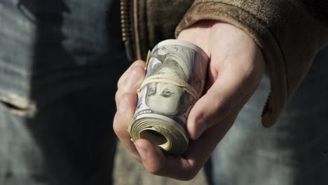 man holds big roll of money, puts it away, outside, sunny day, closeup