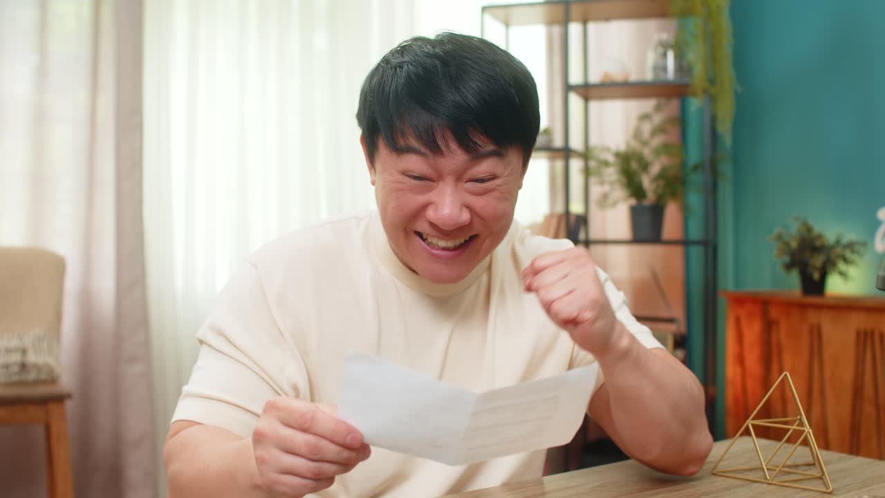 Asian man smiling while reading open letter on paper glad about good news joyful reaction at desk