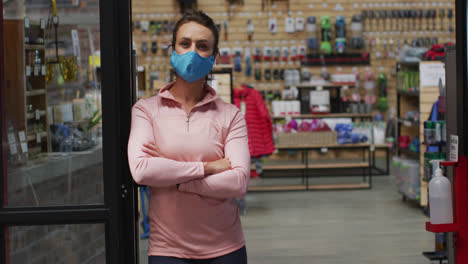 Comerciante-Femenina-Caucásica-Con-Mascarilla-Apoyada-En-La-Puerta-De-La-Tienda-De-Deportes