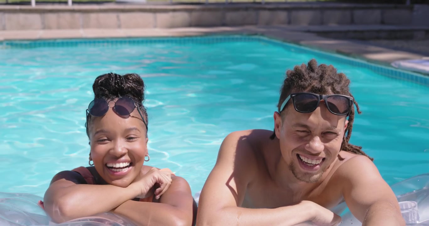 Coppia afroamericana appoggiata a un galleggiante trasparente in piscina a casa, con il sole che luccica, mentre si aggiustano gli occhiali da sole e flirtano