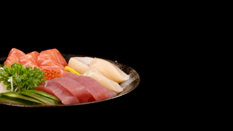 a plate of assorted sashimi, including salmon, tuna, scallop, and salmon roe, rotates smoothly against a black background under bright studio lighting