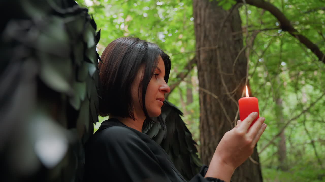 Side view of woman dressed in black with large dark wings holding burning red candle in forest, surrounded by soft green light, symbolizing meditation, mystery, and spiritual ritual in nature setting