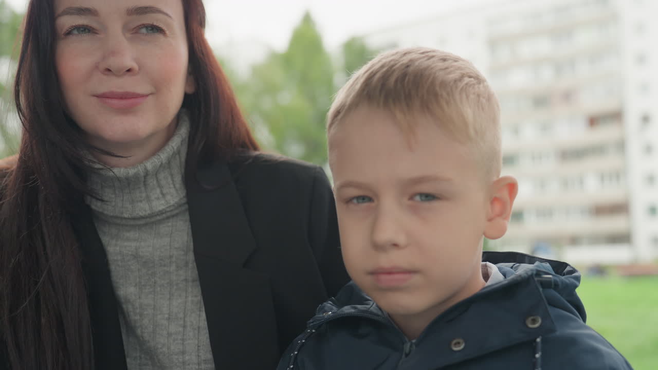 Madre e figlio si preparano per la scuola in un parco residenziale, donna con dolcevita sistema la giacca del ragazzo mentre i condomini sfocano sullo sfondo, luce soffusa e nuvolosa, atmosfera mattutina calma, emozionante