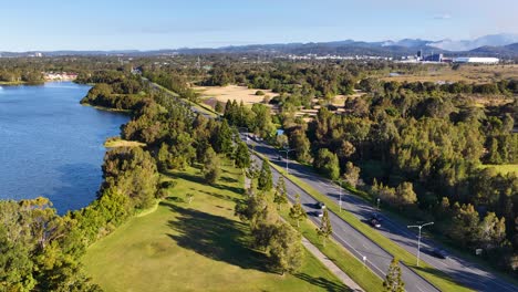 drone footage glides above a tree-lined road running parallel to a river in gold coast, australia, under clear afternoon sunlight with distant mountains visible