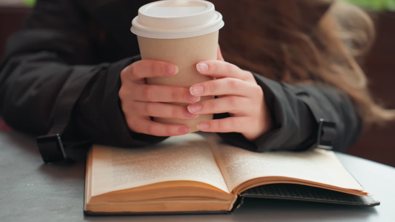 mãos segurando um café sobre um livro, uma jovem pausa a caminhada para tomar um gole de café enquanto lê um livro de bolso em uma mesa aconchegante de um café, usando uma jaqueta escura, luz natural, close-up nas mãos e páginas abertas, ambiente acolhedor