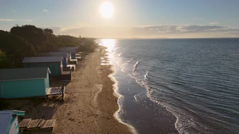 Drone glides above sandy beach and vibrant bathing boxes at sunrise, with calm sea reflections