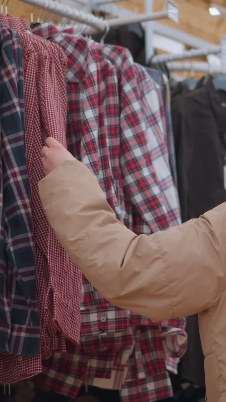 se ve a una chica con una chaqueta marrón y pantalones negros examinando de cerca y tocando la ropa colgada en un estante en una tienda. un momento de compras mientras navega a través de una selección de camisas a cuadros