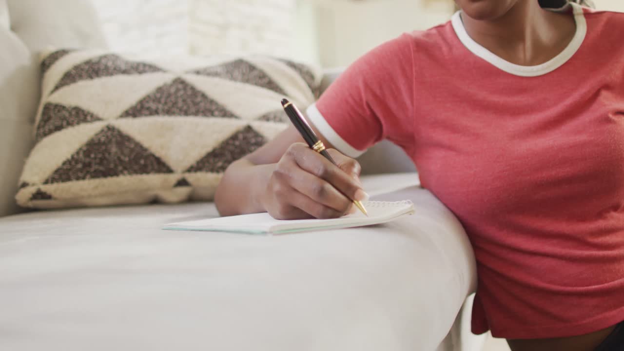 Video of midsection of african american woman sitting on floor, using smartphone and making notes