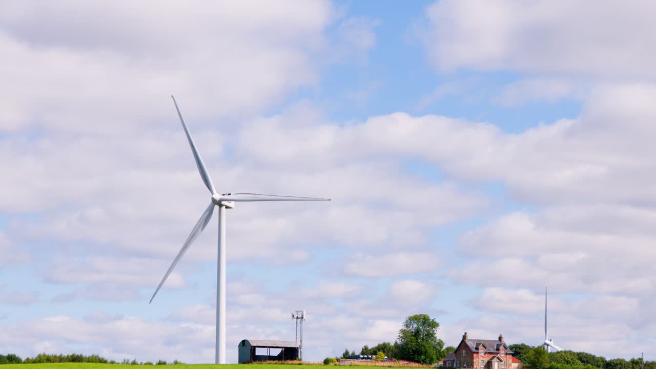 A large wind turbine spins steadily above a green field with grazing cattle and rural houses under bright daylight, captured in a smooth wide shot