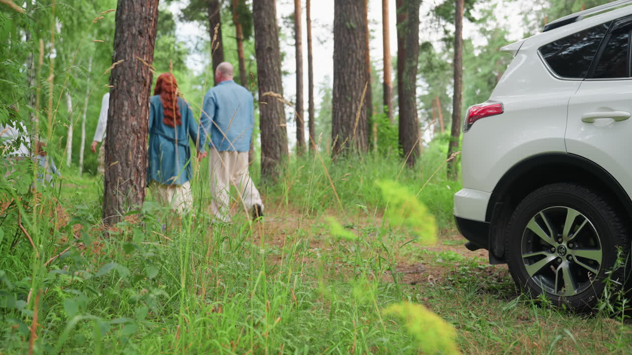 Young couple strolls into peaceful forest with kids holding hands, walking away from parked white car under green trees, sunlight filtering through leaves, warm family outing full of love