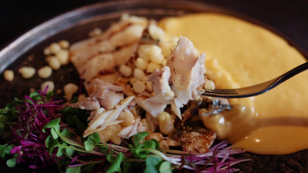 Tampilan dekat hidangan ikan gourmet dengan saus krim, kacang pinus, dan microgreens berwarna-warni.