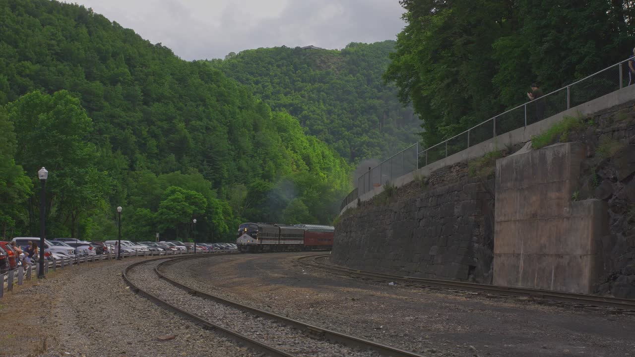 A picturesque view captures train tracks meandering through vibrant green hills, with a parking lot bustling with cars nearby. Nature and transportation blend harmoniously in this serene landscape.
