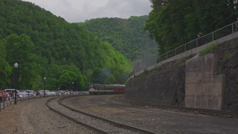 a picturesque view captures train tracks meandering through vibrant green hills, with a parking lot bustling with cars nearby. nature and transportation blend harmoniously in this serene landscape.