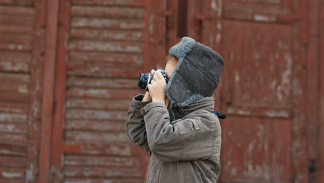 lindo niño pelirrojo con sombrero de aviador tomando fotos con una cámara antigua al aire libre en un fondo de pared de madera