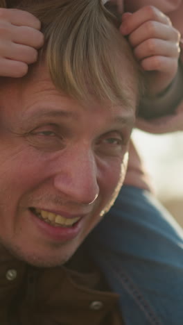 el padre alegre sonríe mientras su niñita, sentada en sus hombros, arregla juguetónmente su cabello en la cálida luz del atardecer