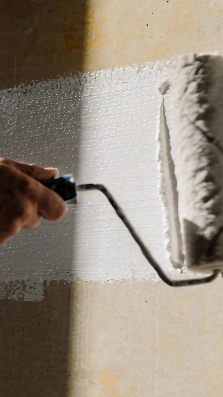 Vertical video: Placing roller at top, hand pulling down on beige wall at home, painting wall white